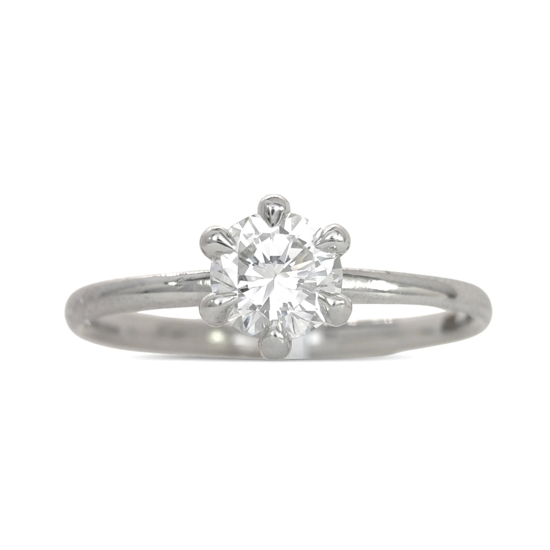 A detailed angled shot of the platinum solitaire ring, focusing on the intricate six-prong mounting and the diamond’s fiery facets. The minimalist design and high polish of the band are highlighted against a stark white surface