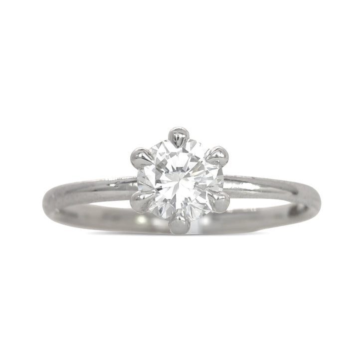 A detailed angled shot of the platinum solitaire ring, focusing on the intricate six-prong mounting and the diamond’s fiery facets. The minimalist design and high polish of the band are highlighted against a stark white surface