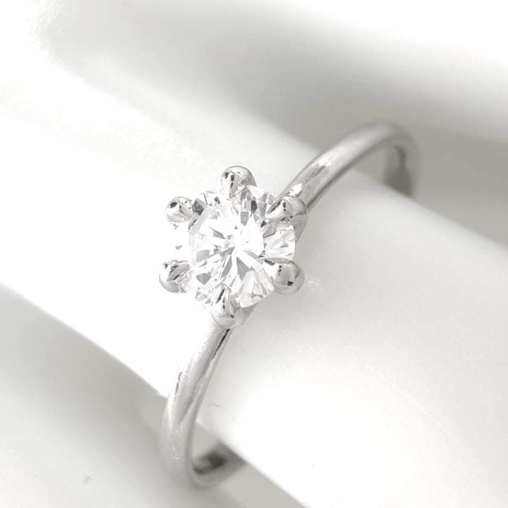 A detailed close-up highlighting the intricate facets of the round diamond and the precision of the six-prong mounting. The polished platinum band reflects light beautifully, set against a minimalist white surface