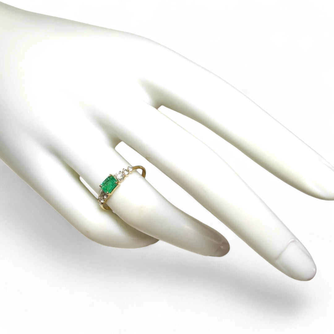 Full view of a womenâs 18K yellow gold engagement ring with a central emerald and diamond accents worn on a mannequin hand
