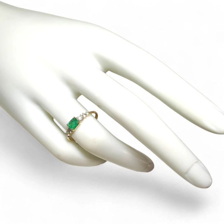 Full view of a womenâs 18K yellow gold engagement ring with a central emerald and diamond accents worn on a mannequin hand