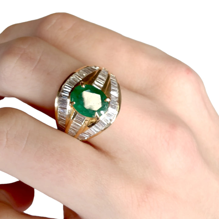 Top view of a woman's hand wearing a lavish 22K gold cocktail ring with a prominent oval emerald and triple bands of diamond baguettes, exemplifying opulence and style.
