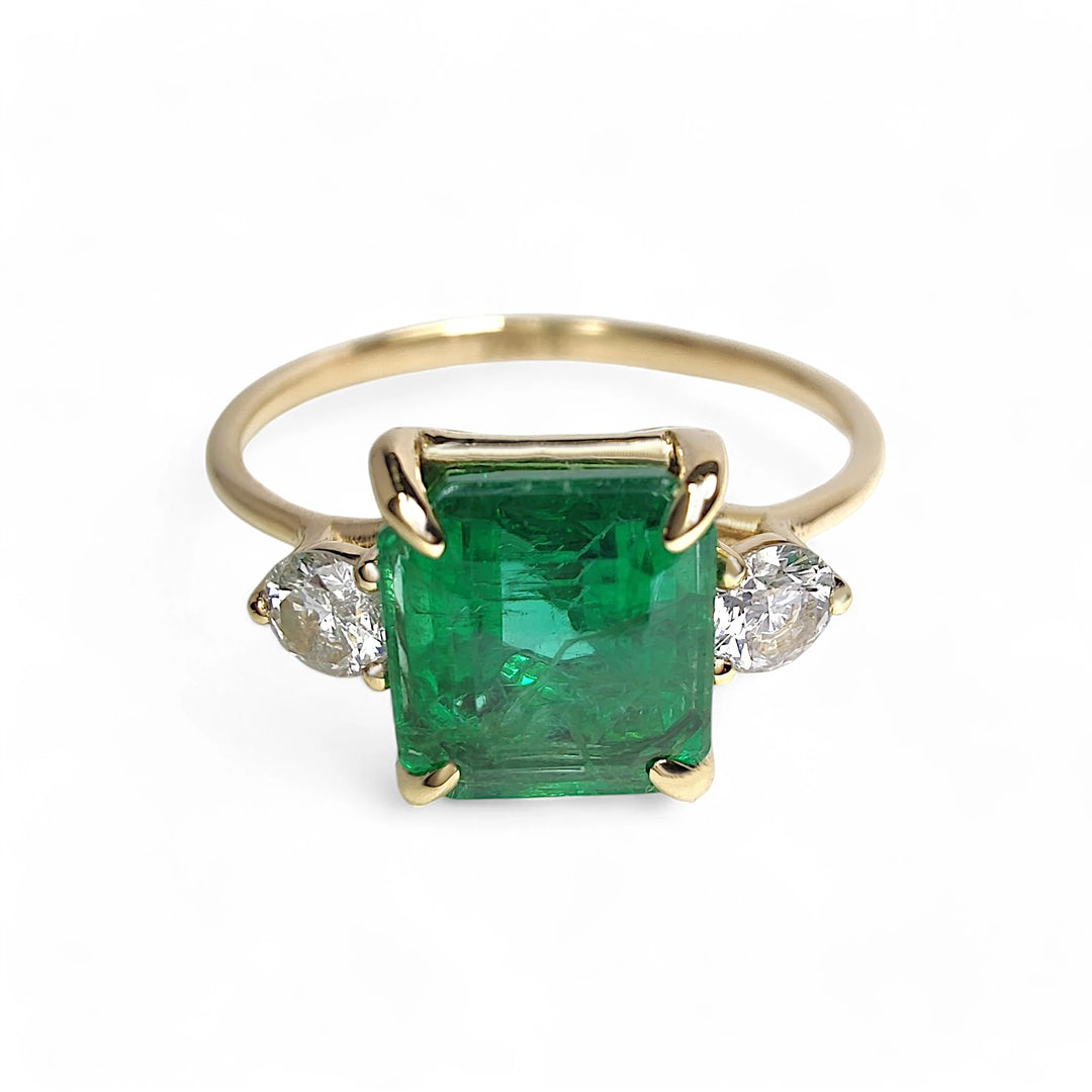 Front view of a womenβs 18K yellow gold engagement ring featuring a central emerald and two round diamonds in a three-stone design