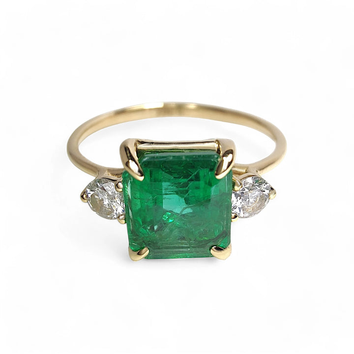 Front view of a womenβs 18K yellow gold engagement ring featuring a central emerald and two round diamonds in a three-stone design