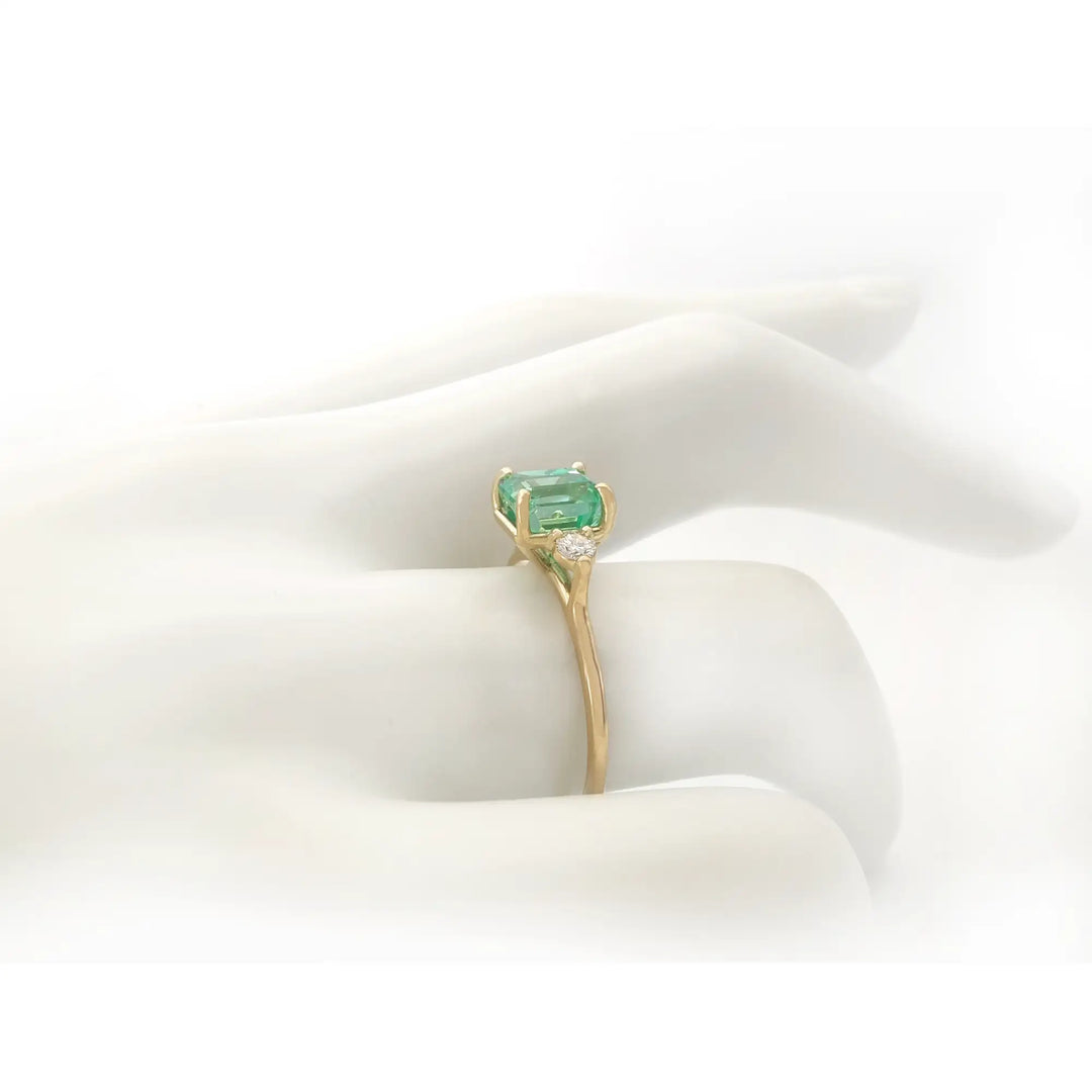 Side view of a three-stone 18K yellow gold engagement ring with an emerald and diamonds displayed on a mannequin hand