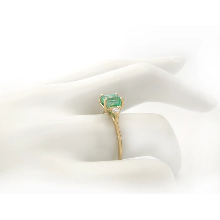 Side view of a three-stone 18K yellow gold engagement ring with an emerald and diamonds displayed on a mannequin hand