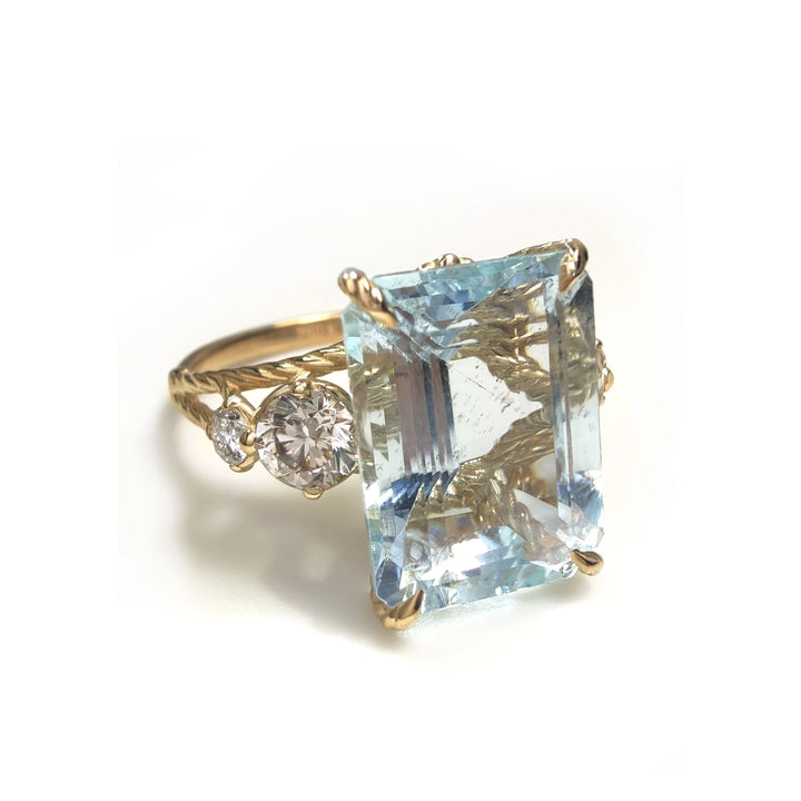 Horizontal perspective view of a women’s 18K yellow gold cocktail ring featuring a large emerald-cut aquamarine, round brilliant-cut diamonds, and a twisted band