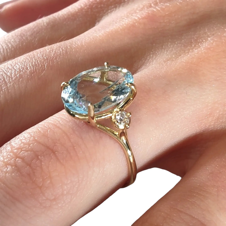 Close-up detail of a three-stone engagement ring in 18K yellow gold with an oval aquamarine and diamond accents displayed on a model’s hand