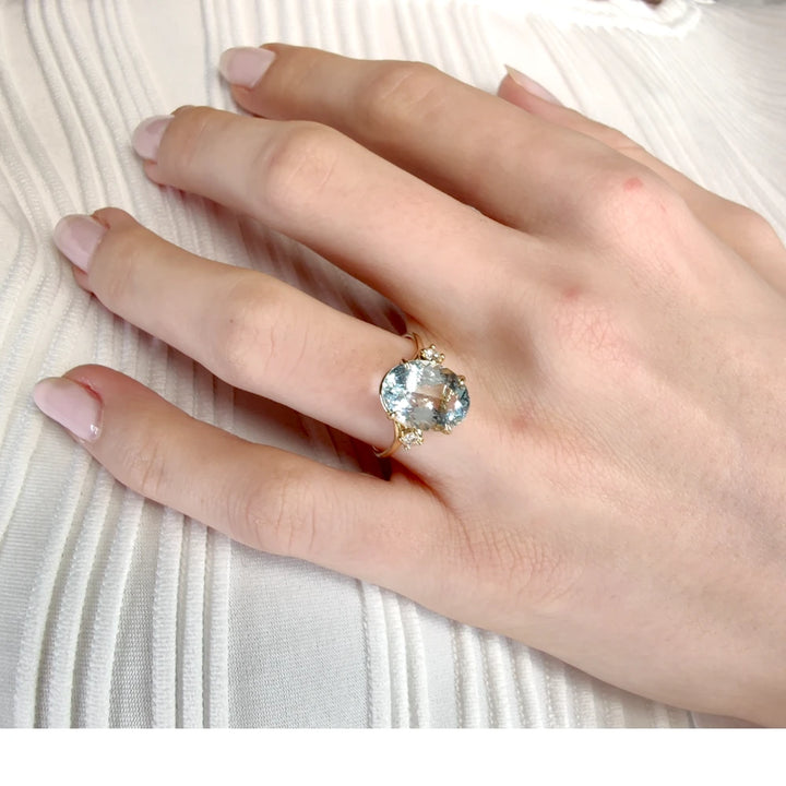 Beautiful three-stone engagement  ring in 18K yellow gold featuring an oval aquamarine and diamonds, worn on a model’s hand in a graceful pose