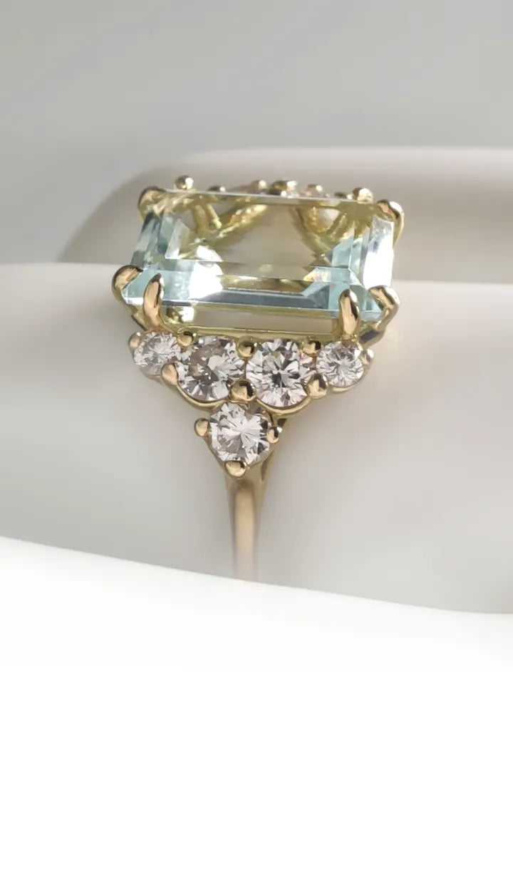 "18K yellow solid gold ring, contemporary style, for women, featuring a large emerald-cut central aquamarine and 5 brilliant-cut diamonds on each side. The ring is displayed on a mannequin hand, close-up lateral view. The white background highlights the light blue of the aquamarine