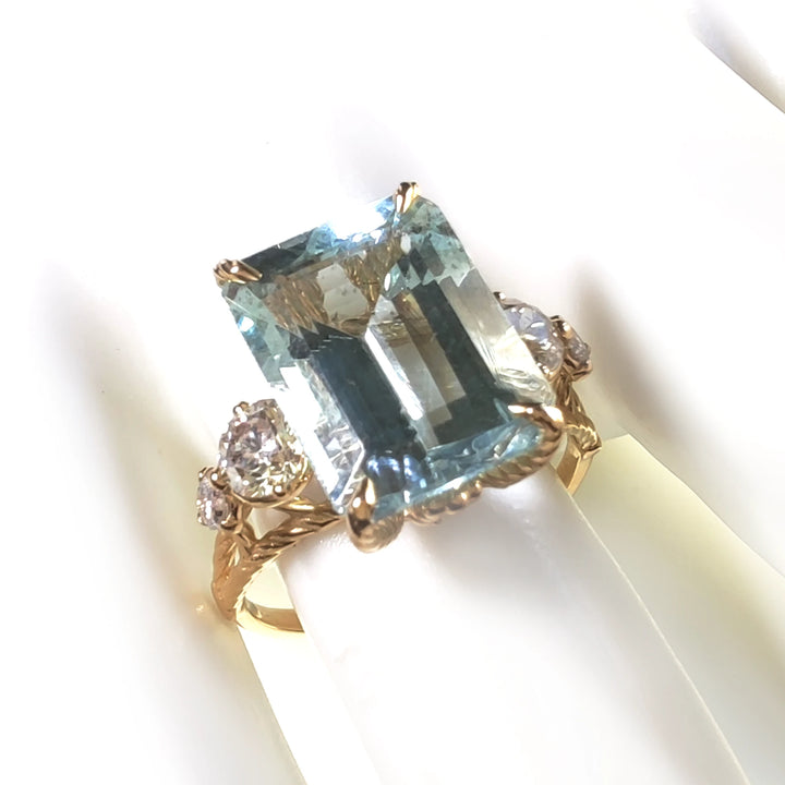 Top view of a 18K yellow gold cocktail ring featuring a large aquamarine centerpiece and diamond accents displayed on a mannequin finger