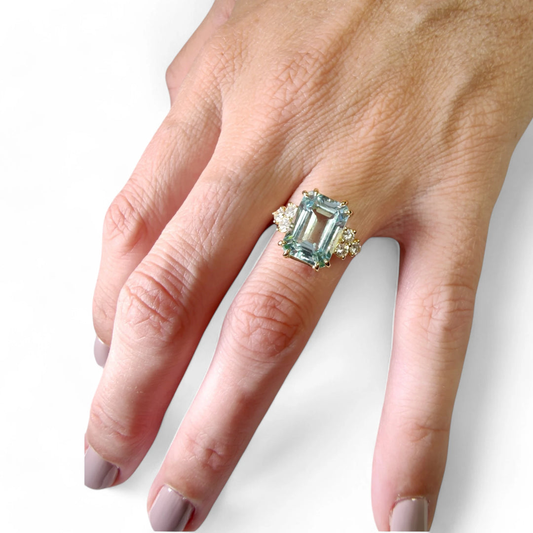 Full hand view of a model wearing a handcrafted 18K yellow gold engagement ring, showcasing a large emerald-cut aquamarine and six diamonds, emphasizing the contemporary design