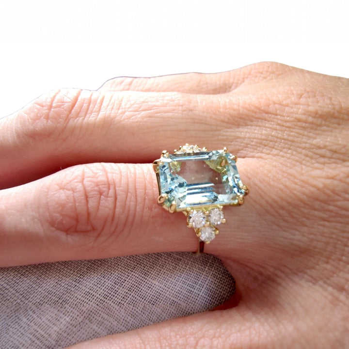 Close-up of a woman's hand displaying an 18K yellow gold engagement ring with a large emerald-cut aquamarine and diamond accents, highlighting the ring's luxurious and handmade details.