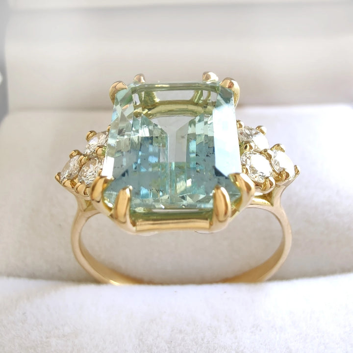 Top view of a jewelry box containing a contemporary 18K yellow gold engagement ring with a large emerald-cut aquamarine and cathedral-style diamonds, showcasing the elegant presentation