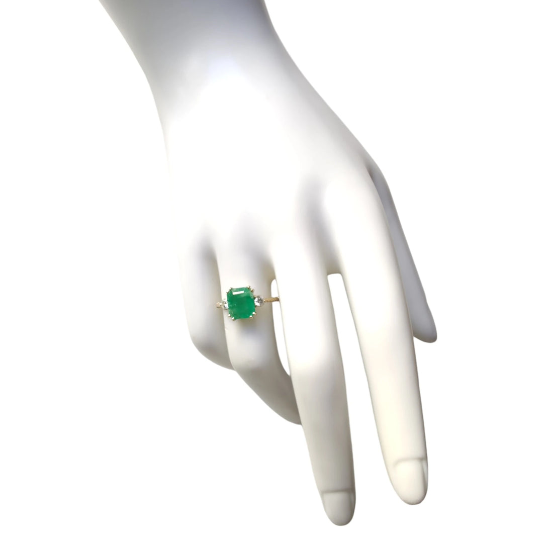 View of an 18K yellow gold three-stone engagement ring on a mannequin hand, showcasing a central emerald flanked by diamonds, perfect to buy for a Christmas gift