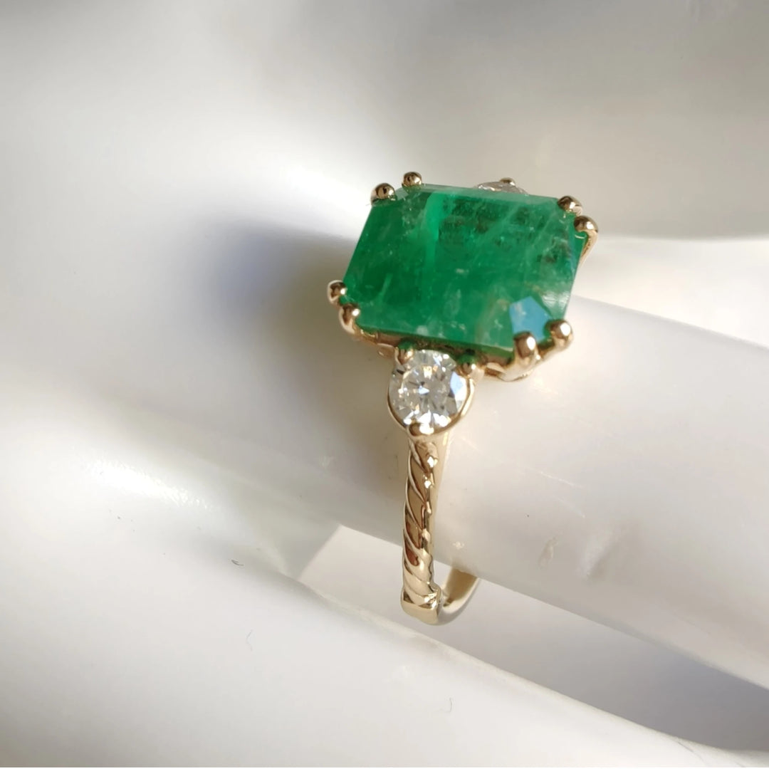 Close-up view of a three-stone emerald engagement ring in 18K yellow gold, displayed on a mannequin hand, highlighting the ring’s exquisite details and craftsmanship