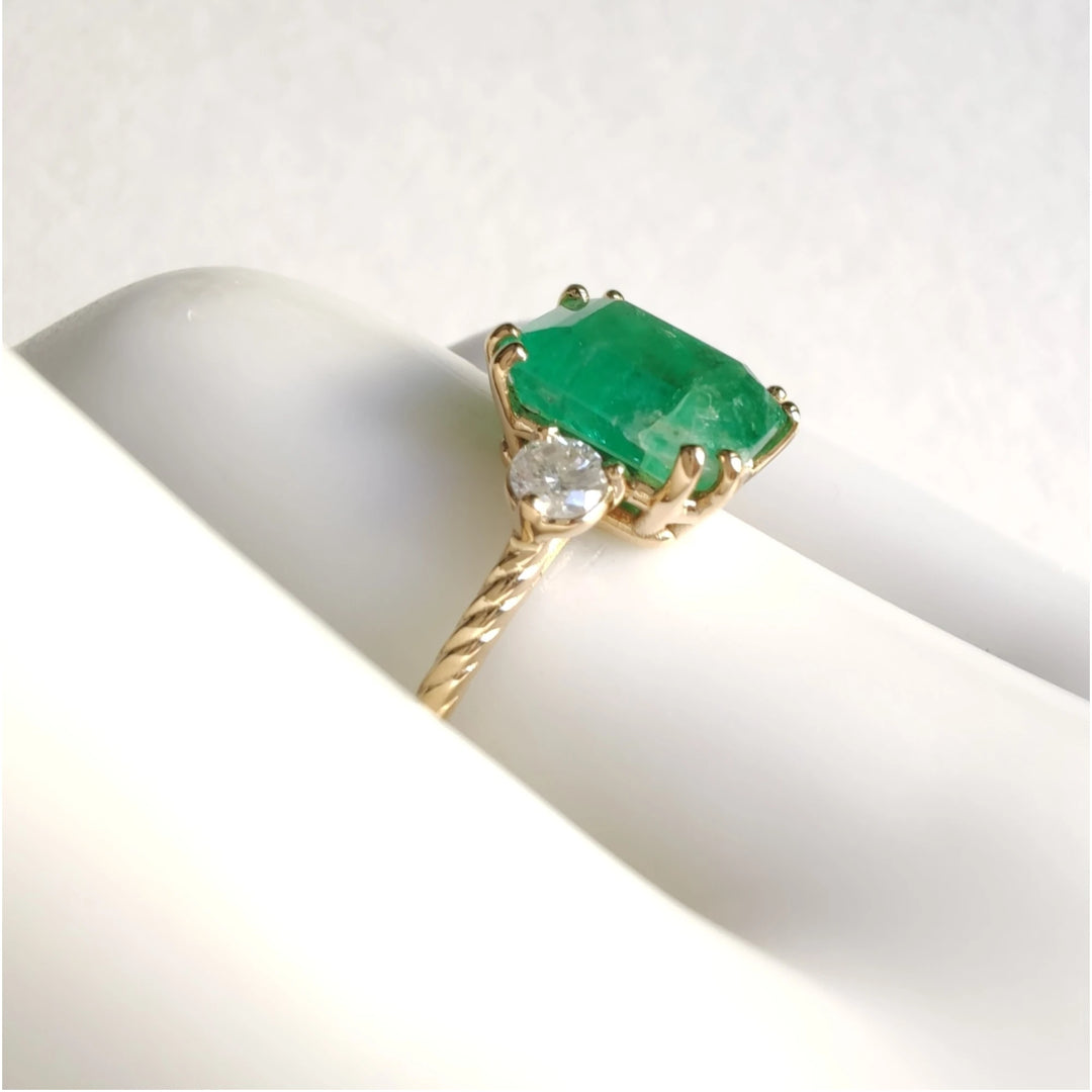 Side view of an 18K yellow gold engagement ring on a mannequin hand, featuring a central emerald and diamonds, ideal for those looking to buy a timeless engagement ring