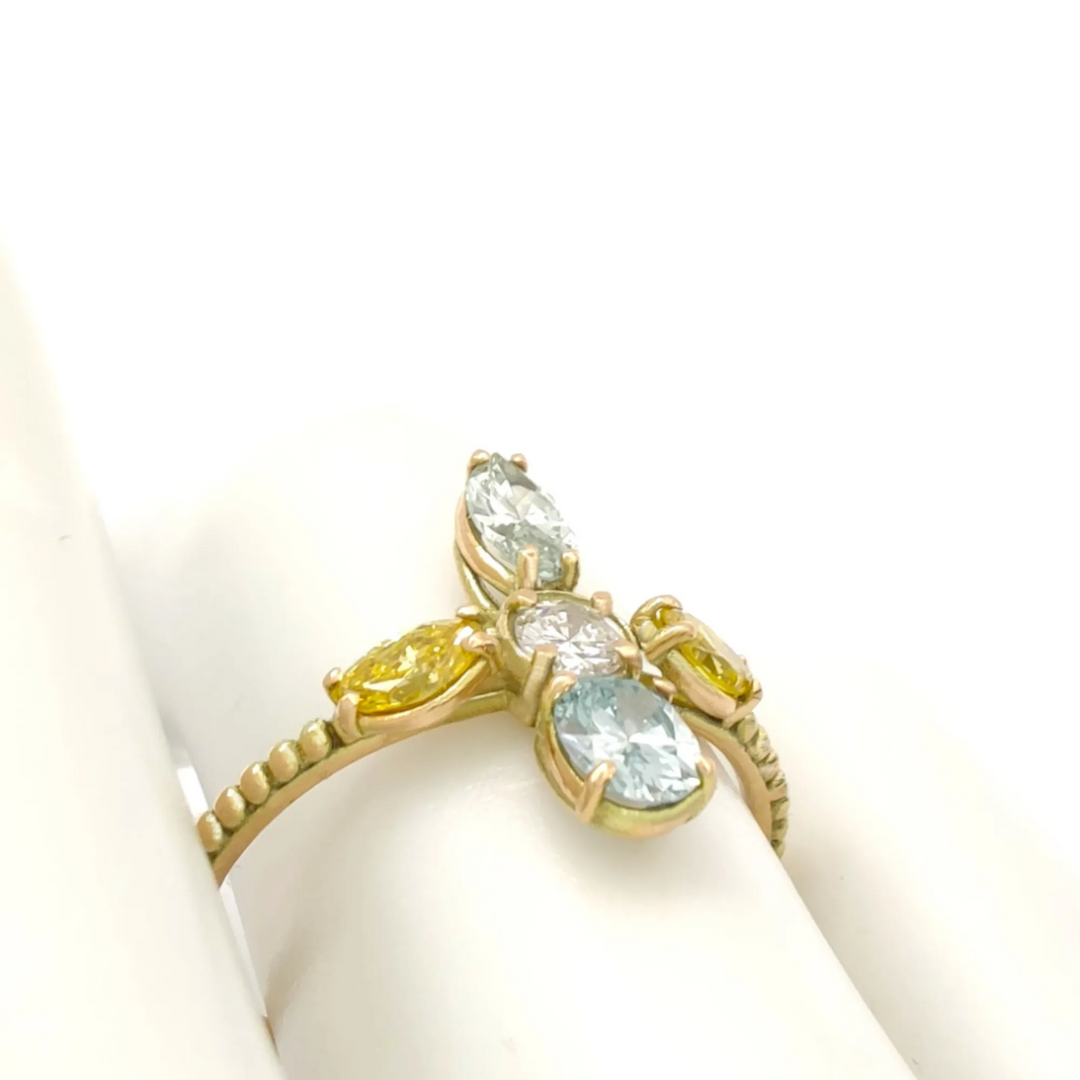 14K yellow gold engagement ring on a mannequin hand featuring a cross design with miligrain details, a central round brilliant diamond, and surrounding fancy colored diamonds, ideal for a unique engagement proposal