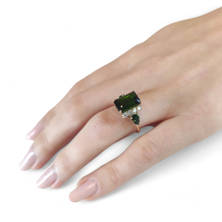 Full hand view of a model wearing a 14K yellow gold minimalist ring, showcasing an emerald-cut dark green tourmaline with brilliant diamonds and pear-shaped side tourmalines, ideal for modern jewelry lovers seeking a unique styl