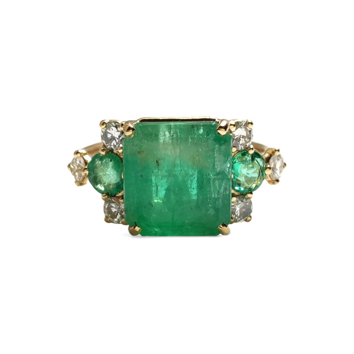 Front view of a 18K yellow gold engagement ring with a central emerald cut emerald, two round emeralds, and three brilliant cut diamonds