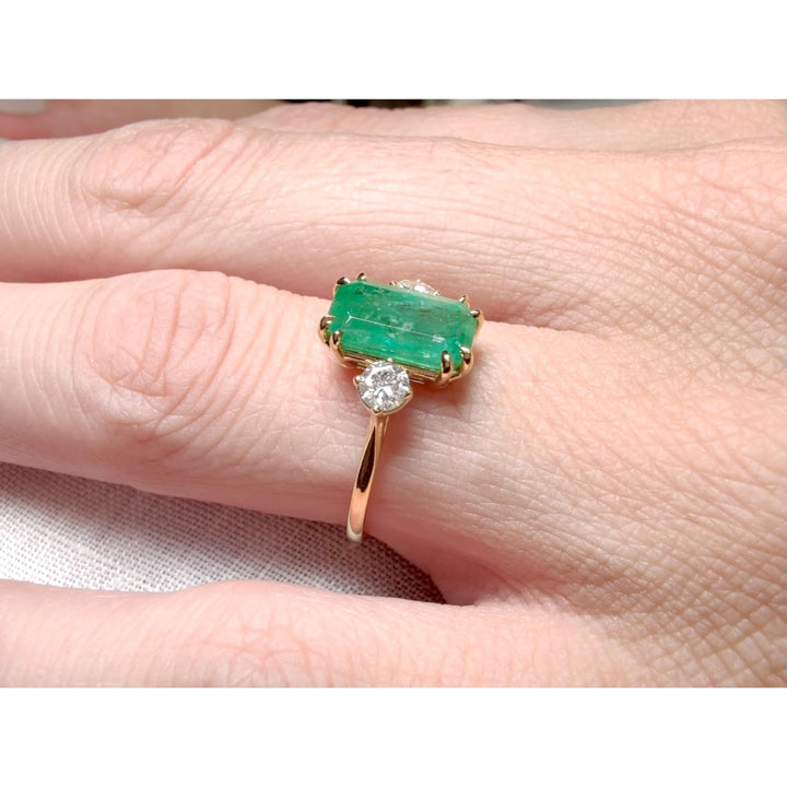 Close-up view of a women’s three-stone engagement ring in 18K yellow gold with an emerald centerpiece and diamonds displayed on a model’s hand