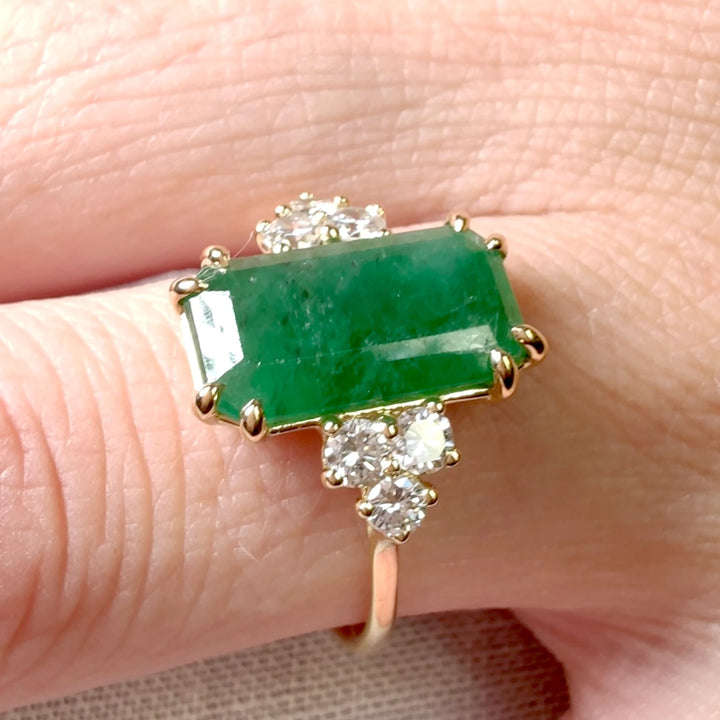 Detailed close-up of a fine jewelry engagement ring with a central emerald and diamonds set in 18K yellow gold worn on a model’s finger