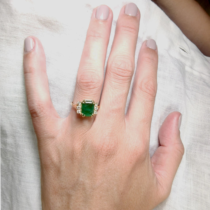 Front view of a women’s 18K yellow gold engagement ring with emerald and diamonds displayed on a model’s hand, perfect for proposals or gifting occasions