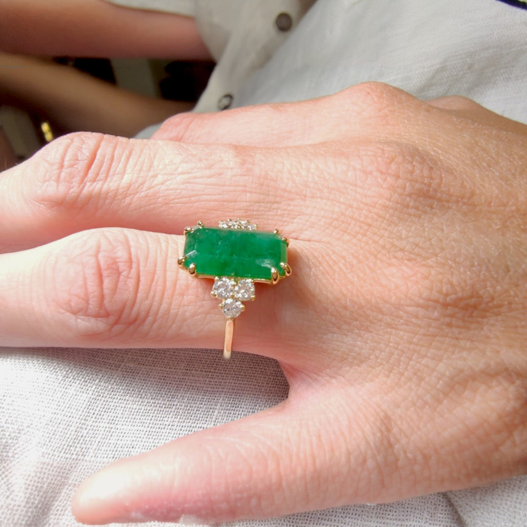 Close-up side view of a women’s 18K yellow gold engagement ring with emerald and diamond accents displayed on a model’s hand