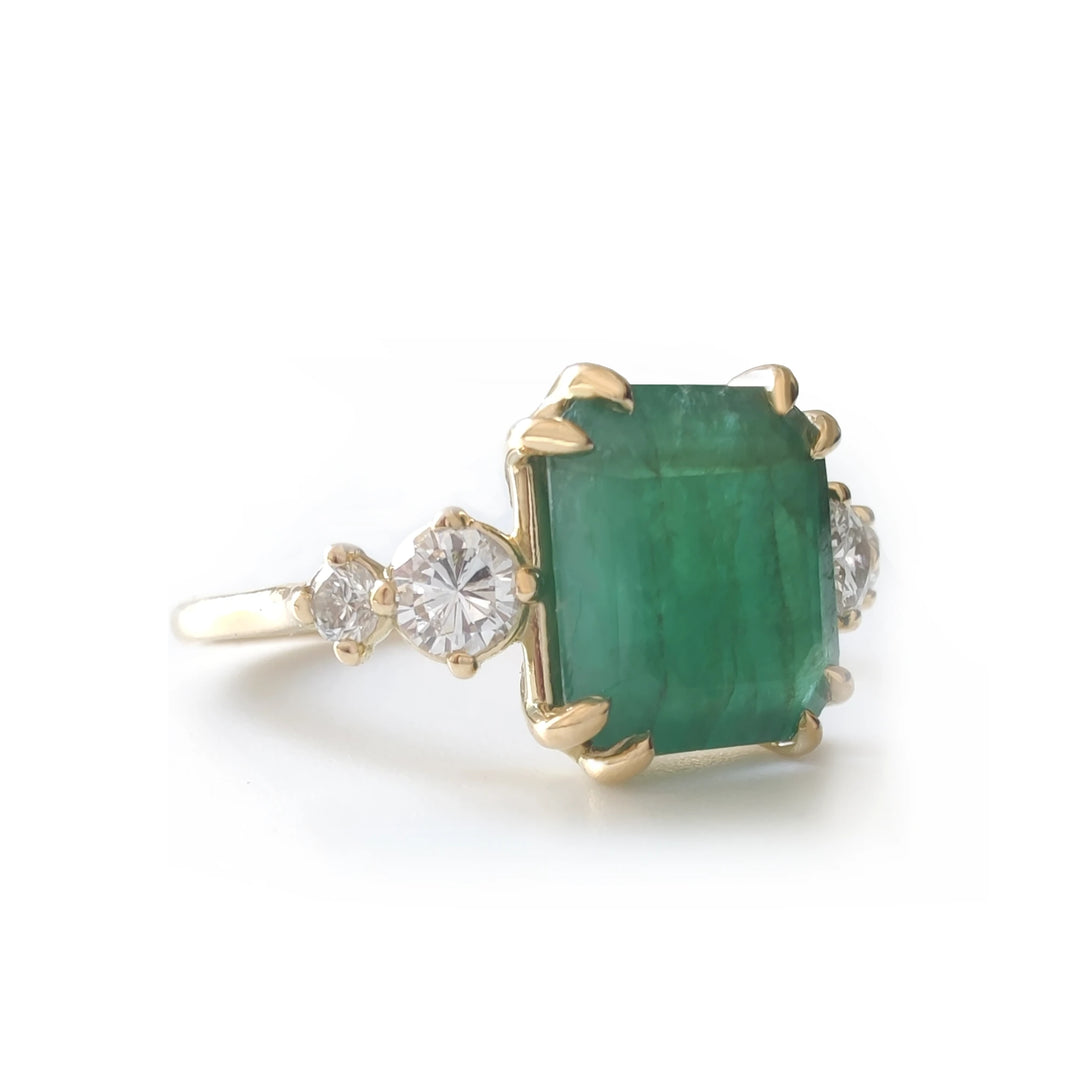 Left side view of a timeless 18K yellow gold engagement ring with a central emerald cut emerald and round diamonds on each side