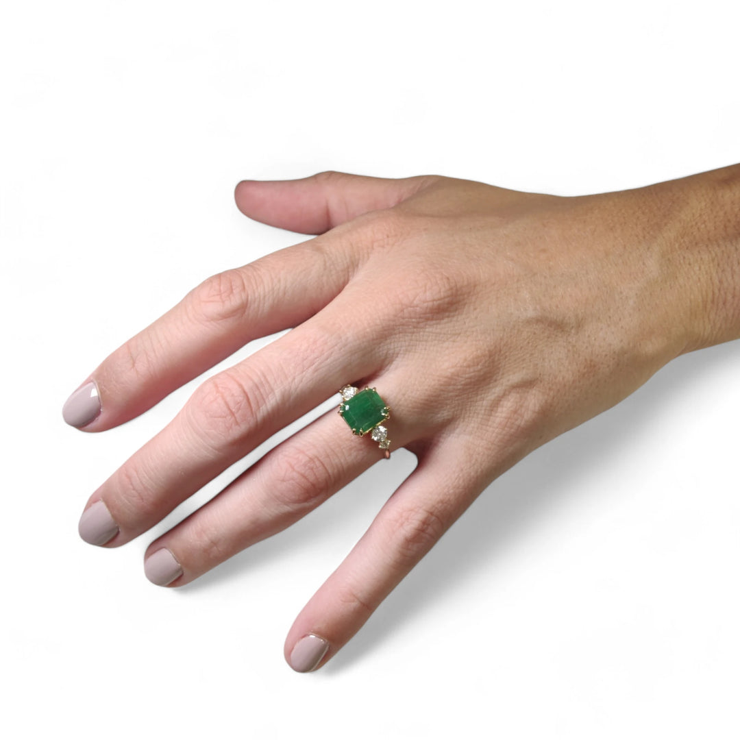 Elegant 18K yellow gold engagement ring with a central emerald cut emerald and diamond accents worn on a woman’s hand