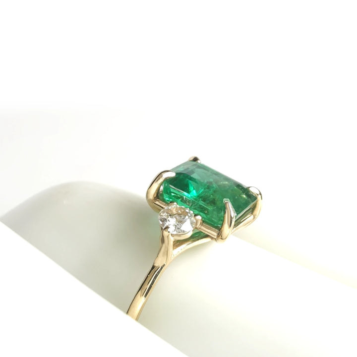 Side detail view of a 18K yellow gold three-stone ring with a central emerald and round diamond accents displayed on a mannequin hand