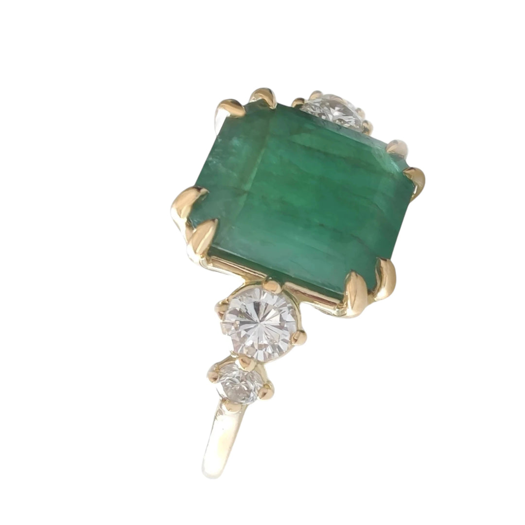 Perspective view of a women’s 18K yellow gold engagement ring featuring a central emerald cut emerald and two round diamonds on each side in a tapered design