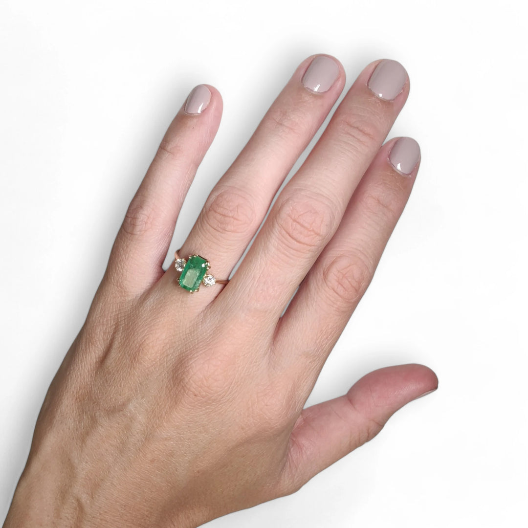 Elegant 18K yellow gold three-stone engagement ring with a central emerald and diamonds worn by a female model, ideal for proposals or holiday gifting