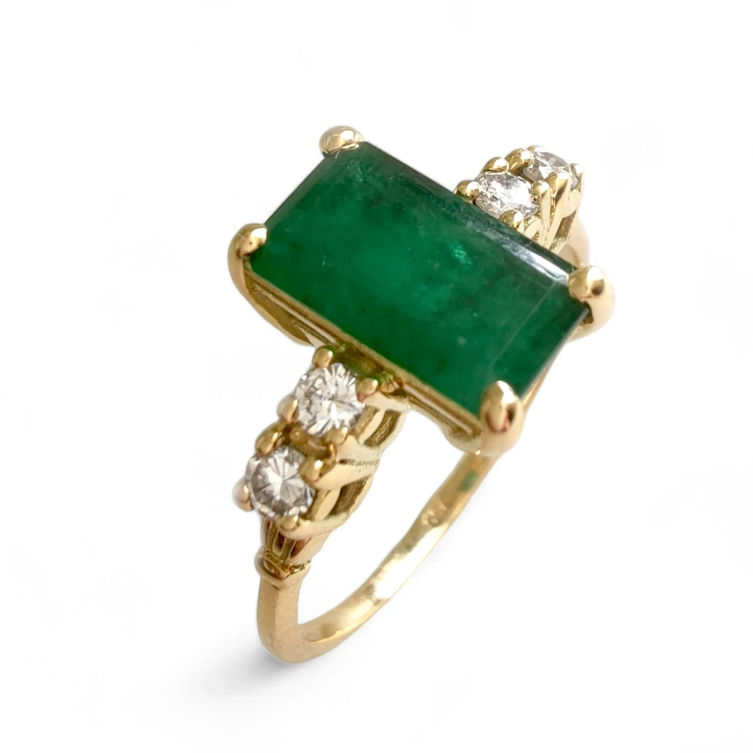 Vertical perspective view from above of an 18K yellow gold engagement ring featuring a central emerald-cut emerald and two brilliant-cut diamonds on each side in a classic vintage design