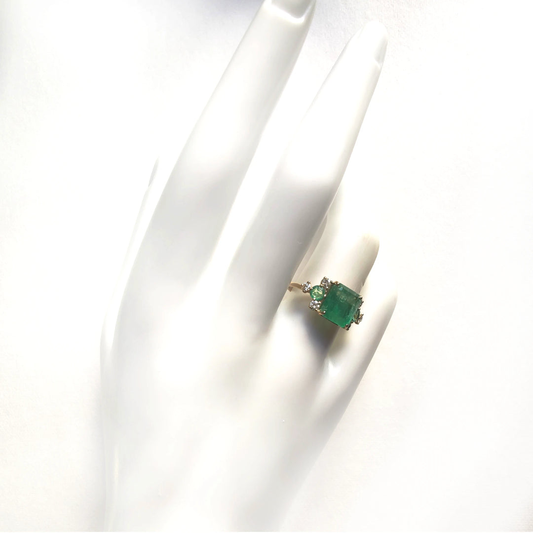 Elegant 18K yellow gold engagement ring featuring an emerald cut emerald, round emeralds, and brilliant diamonds displayed on a mannequin hand