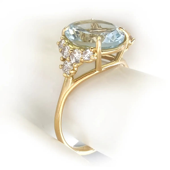 Close-up view of the left side of a handcrafted 18K yellow gold engagement ring with a genuine oval aquamarine and brilliant diamonds on a mannequin finger.